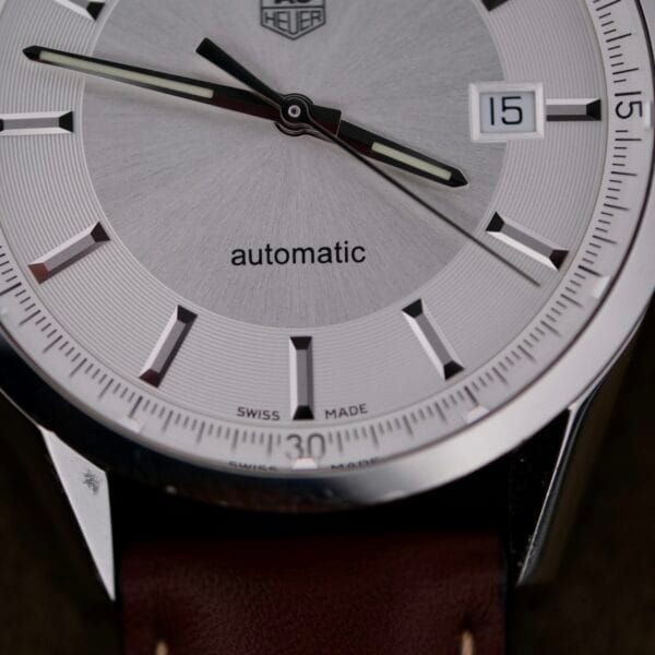 The Tag Heuer Carrera Caliber 5 Automatic Wv211A.Fc6203 features a vintage silver watch face with black and white hour markers and hands, labeled automatic. It has a date display at 3 oclock and comes with a classic brown leather strap.