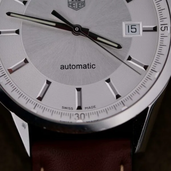 The Tag Heuer Carrera Caliber 5 Automatic Wv211A.Fc6203 features a vintage silver watch face with black and white hour markers and hands, labeled automatic. It has a date display at 3 oclock and comes with a classic brown leather strap.