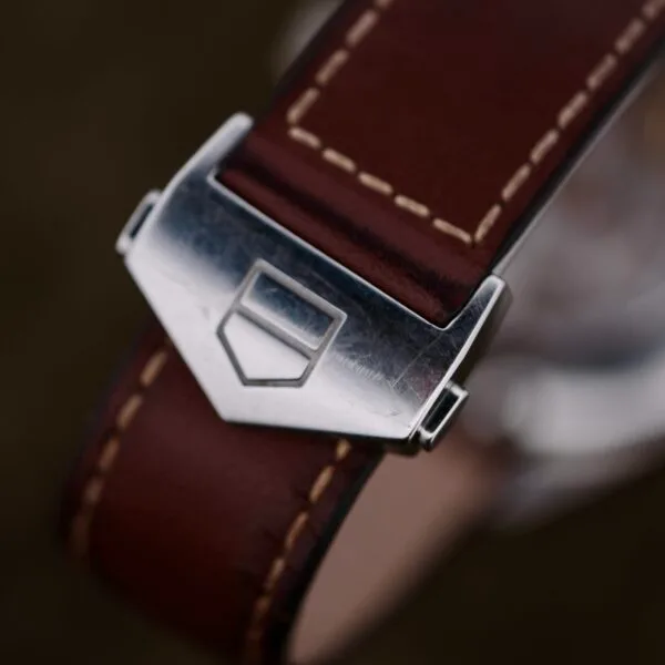 A close-up of the Tag Heuer Carrera Caliber 5 Automatic leather watch strap reveals its vintage brown leather design, white stitching, and silver clasp with an engraved emblem, set against a softly blurred background that highlights its timeless appeal.