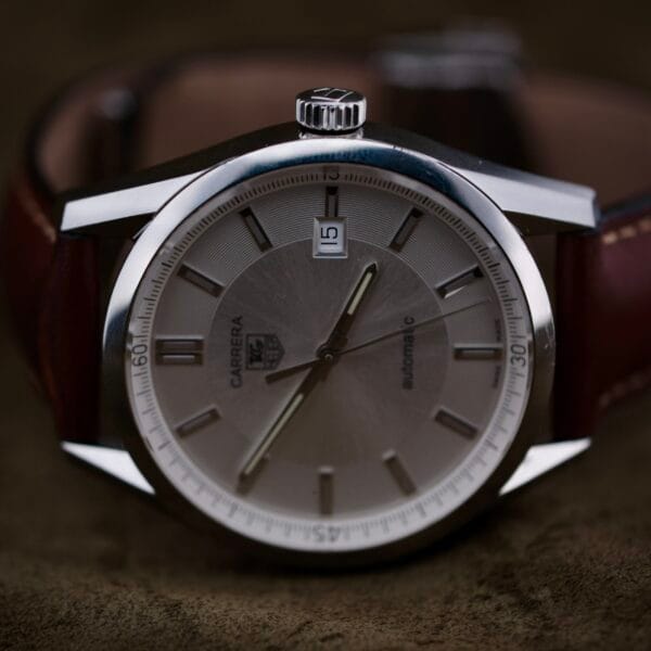 A close-up of a Tag Heuer Carrera Caliber 5 Automatic Wv211A.Fc6203 wristwatch features a vintage silver face, brown leather strap, date window at 3 oclock, and silver hour markers on a soft, dark background.
