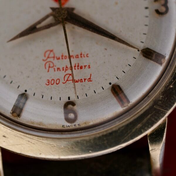A close-up of the Omega Pinpointers 300 Game Dial Wristwatch Automatic reveals vintage metal hands. The dial features Automatic Pinspotters 300 Award in red. Its charming weathered look, with patina and dust, hints at a storied past.
.