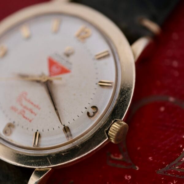 A close-up of the Omega Pinpointers 300 Game Dial Wristwatch Automatic displays a white face with gold-tone numbers and hands. This vintage watch, with its black leather strap, elegantly rests on a textured red surface embellished with gold lettering.