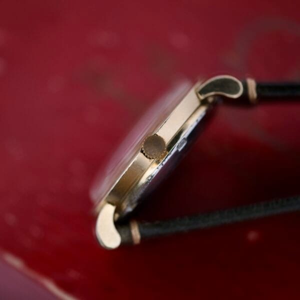 Close-up side view of the Omega Pinpointers 300 Game Dial Wristwatch Automatic, featuring a vintage golden case and black leather strap, set against a blurred red background.