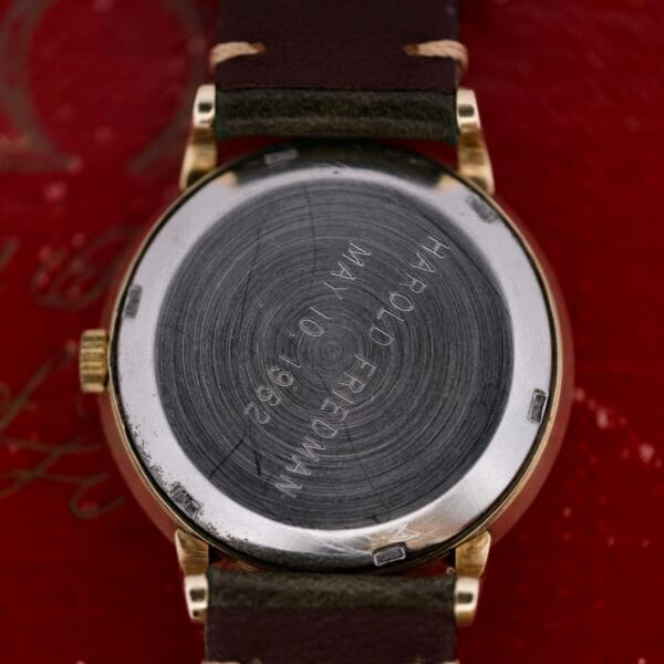 A close-up of the back of an Omega Pinpointers 300 Game Dial Wristwatch Automatic engraved with HAROLD FRIEDMAN MAY 10, 1962. The classic watch, featuring a dark strap, is elegantly placed on a red surface.