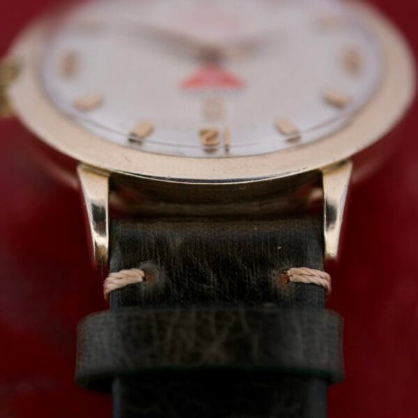 A close-up of the Omega Pinpointers 300 Game Dial Wristwatch Automatic showcases a gold-toned case and textured black leather strap. Gold hour markers and hands shine against a blurred red backdrop, while the slightly worn case adds to its timeless charm.