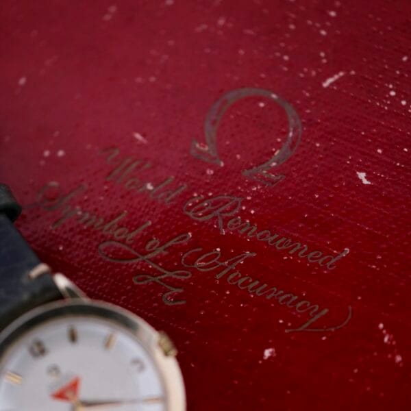 A close-up of an Omega Pinpointers 300 Game Dial Wristwatch Automatic with a dark wristband rests beside a textured red surface, featuring a faded logo and the phrase World Renowned Symbol of Accuracy in cursive.