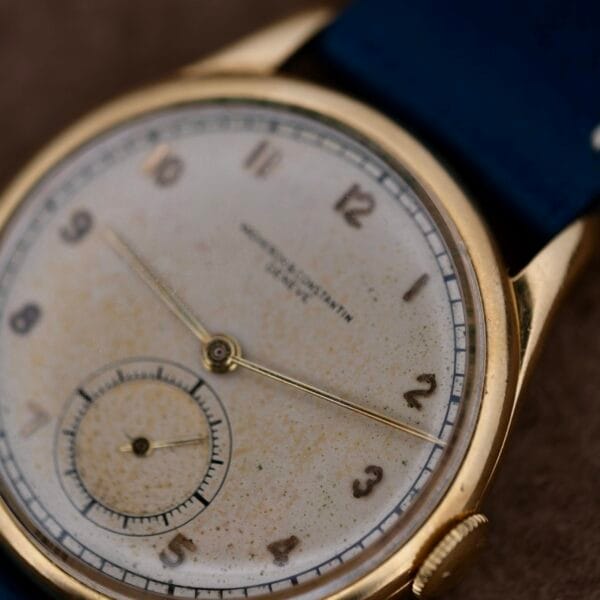 The Vacheron Constantin 18k Gold Caltrava Cal. V453 features a worn, off-white dial with gold-tone Arabic numerals and hands, a small subdial, and a dark blue strap. The elegant 18k gold case completes the timeless design of this classic vintage wristwatch.