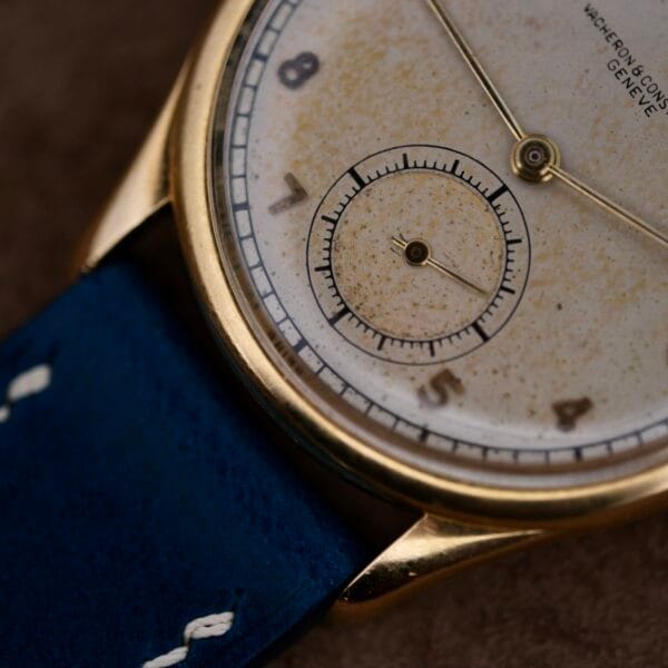 This close-up features a vintage Vacheron Constantin 18k Gold Caltrava Cal. V453 wristwatch, with a weathered dial, Arabic numerals, subdial, and complemented by a blue leather strap with white stitching.