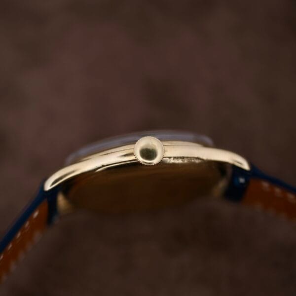 A close-up of the Vacheron Constantin 18k Gold Caltrava Cal. V453 watch crown on a gold case, complemented by blue and brown leather straps. The blurred background accentuates its timeless elegance.