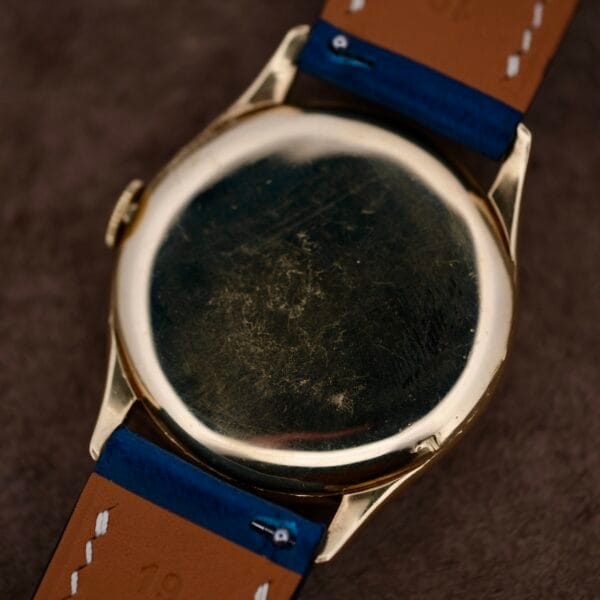 A scratched Vacheron Constantin 18k Gold Caltrava Cal. V453 watch with a leather strap featuring blue and brown sections is focused on its back against a dark, textured background, highlighting its timeless elegance.