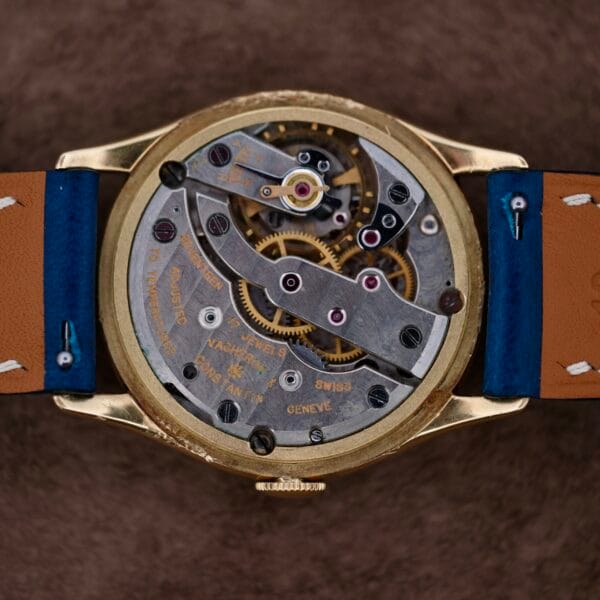 The Vacheron Constantin 18k Gold Caltrava Cal. V453 showcases its intricate mechanical gears through an open back. The elegant watch has a gold casing, a brown leather strap with blue accents, all beautifully arranged on a textured brown surface.