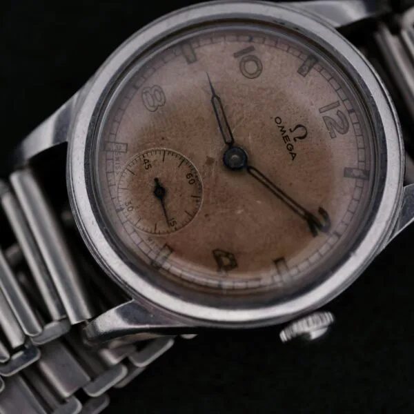 An Omega WWII Era Military Sub Seconds wristwatch with a silver metal strap is on display. It features a patina on its face, Arabic numerals, and a small seconds subdial, with a polished silver case finish.