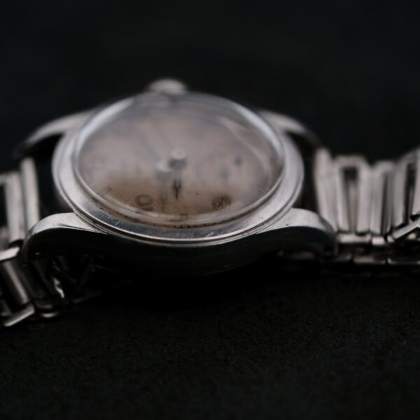 A close-up of an Omega WWII Era Military Sub Seconds wristwatch with a metal band and a scratched, faded dial against a dark background. The visible hands emphasize its timeless charm and aged condition.