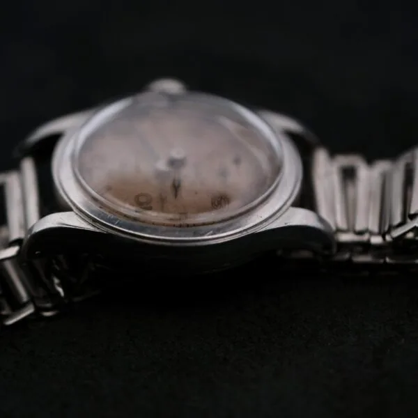 A close-up of an Omega WWII Era Military Sub Seconds wristwatch with a metal band and a scratched, faded dial against a dark background. The visible hands emphasize its timeless charm and aged condition.