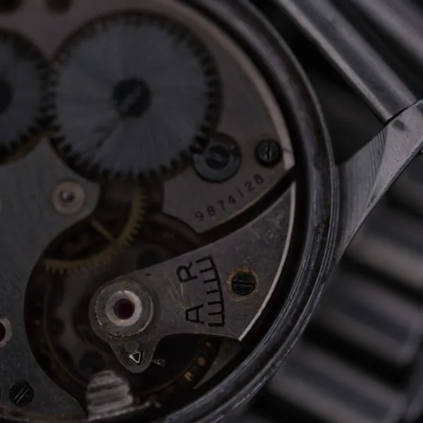 Close-up of an Omega WWII Era Military Sub Seconds watch movement, displaying intricate gears and metal components. The image highlights craftsmanship with a partial view of the vintage mechanism, featuring serial number 9874128 and AR markings on the metal surface.
