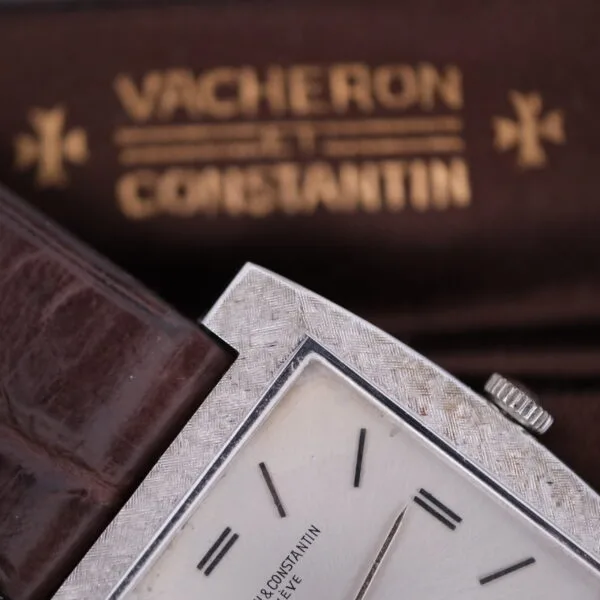 Close-up of a Vacheron Constantin 6761 Cal 1003 watch, featuring a textured silver square case, brown leather strap, dark hands, and index marks. The brown background displays the brands name in gold lettering.