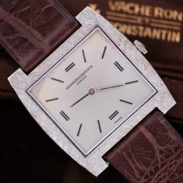 The Vacheron Constantin 6761 Cal 1003 boasts a square design with a textured silver bezel and simple white dial featuring thin black hands and markers, complemented by a brown leather strap. The brand name highlights the dial, packaged elegantly in its branded box.