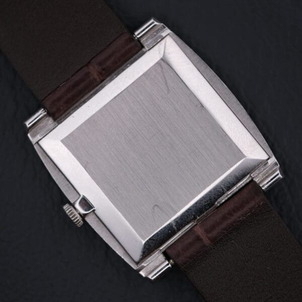 A silver square watch case with a dark brown leather strap is set against a dark background. The blank watch face subtly reflects light, reminiscent of the refined elegance of the Vacheron Constantin 6761 Cal 1003, exuding timeless sophistication.
