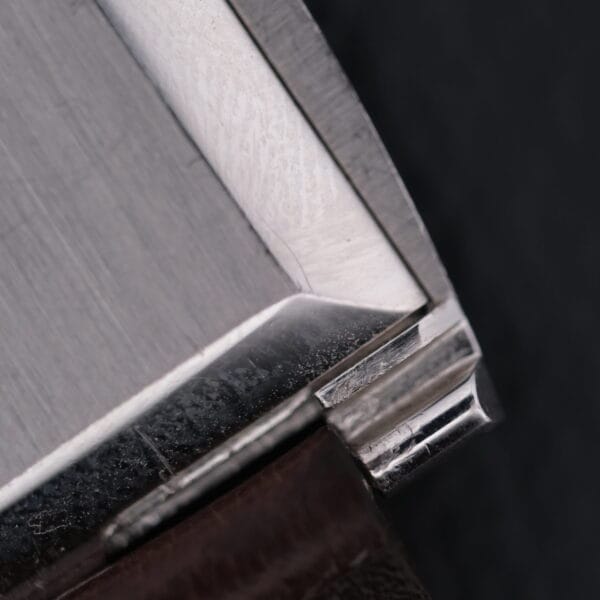 Close-up of an angular, metallic corner of a sleek, brushed steel Vacheron Constantin 6761 Cal 1003 with subtle reflections on its surface against a dark background, highlighting the precision and craftsmanship.