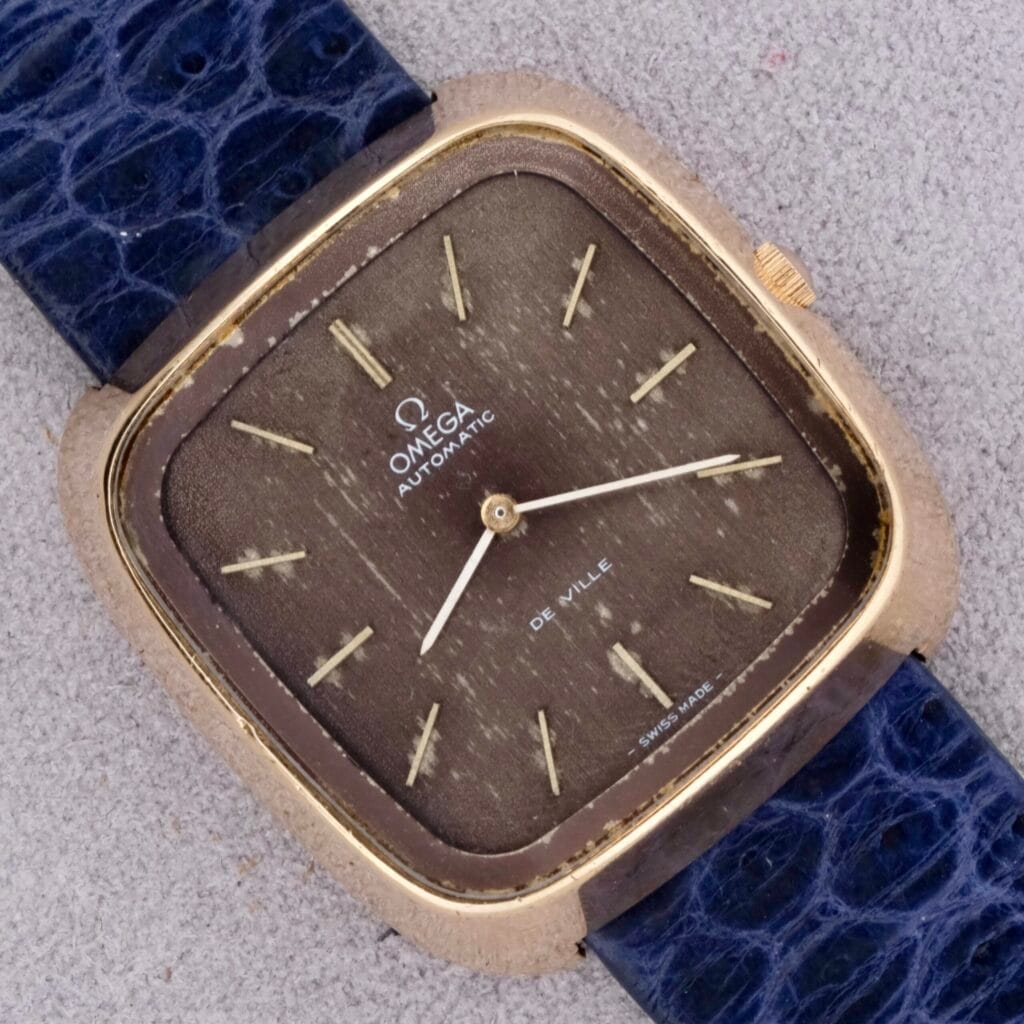 A 1974 Omega Deville Automatic TV Case Ref. 151.0044 with a square gold case and blue textured leather strap rests elegantly on a light surface.