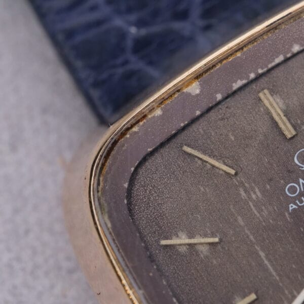 Close-up of a 1974 Omega Deville Automatic TV Case Ref. 151.0044 watch face with a worn surface, gold-tone casing, and hour markers exuding timeless elegance. A textured blue background and light-colored surface enhance the watchs classic charm.