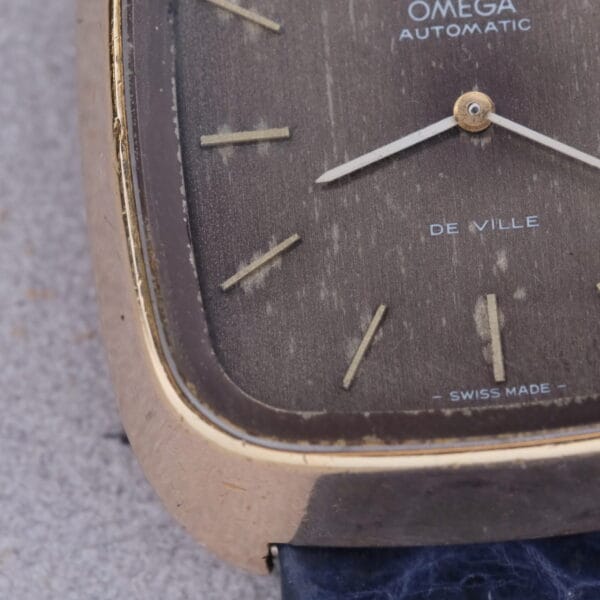 A close-up of the 1974 Omega Deville Automatic TV Case Ref. 151.0044 reveals its vintage dial within a gold-tone rectangular case. Simple hour markers and a date display grace its textured surface, embodying timeless elegance.