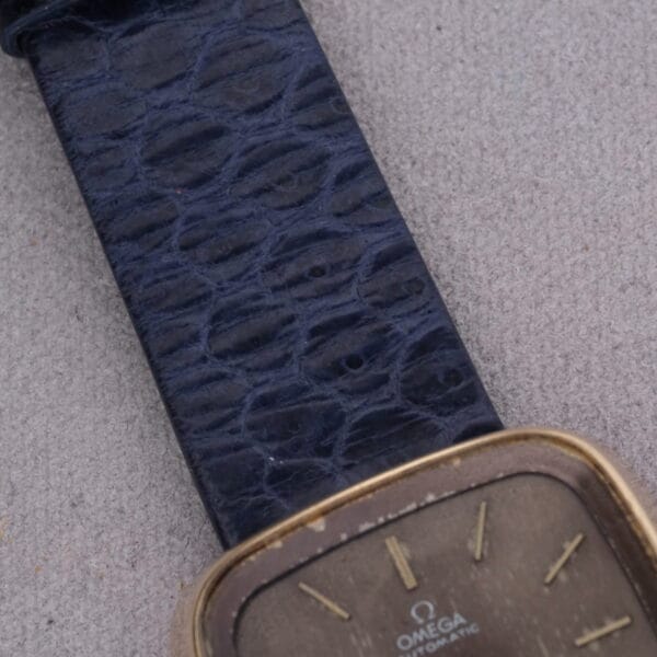 A close-up of the 1974 Omega Deville Automatic TV Case Ref. 151.0044 showing a dark textured leather strap and gold-toned rectangular face with simple hour markers and OMEGA AUTOMATIC on the dial, elegantly resting against a light gray fabric background.