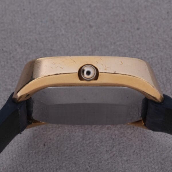A close-up of the 1974 Omega Deville Automatic TV Case Ref. 151.0044s gold-toned case, side view, with its centered crown. The black strap elegantly contrasts against the textured gray surface beneath.