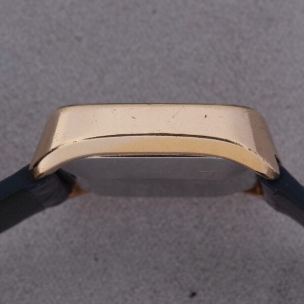 Side view of a 1974 Omega Deville Automatic TV Case Ref. 151.0044 with a gold-toned case and black leather strap, highlighting the curved edge against a gray background.