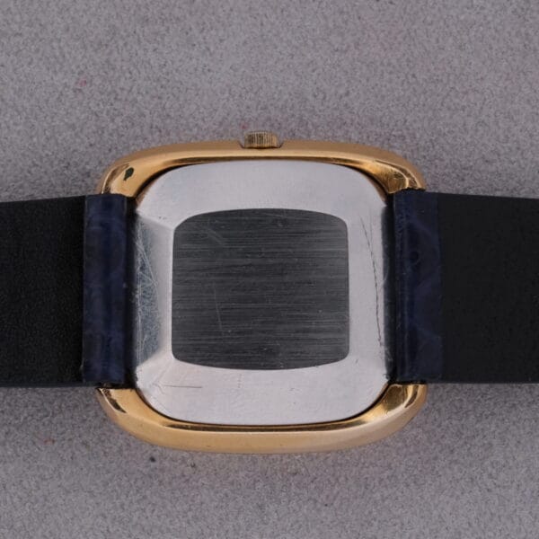 The image features the back of a 1974 Omega Deville Automatic TV Case Ref. 151.0044, displaying its rectangular metal case with a gold-tone edge and an attached black leather strap, along with slight scratches adding character to the watchs surface.