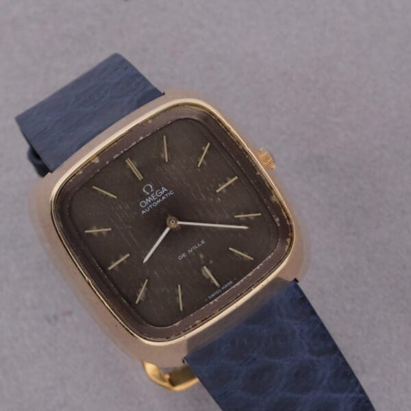 Close-up of a 1974 Omega Deville Automatic TV Case Ref. 151.0044 wristwatch, featuring a square gold-toned bezel, textured dark brown dial, blue textured strap, and gold hour markers. The watch face displays the Omega logo and Automatic text.
