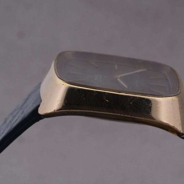 A close-up side view of the 1974 Omega Deville Automatic TV Case Ref. 151.0044 showcases its elegant rectangular black face and gold-colored frame, with a dark strap contrasting against a neutral background, highlighting its timeless sophistication.