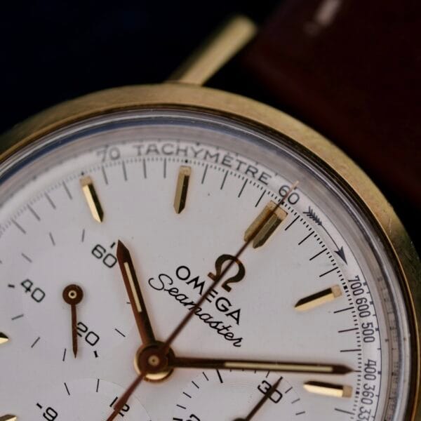 Close-up of the Omega Seamaster Chronograph Ref. 105.005 321 Caliber, featuring a gold case, white dial, and chronograph scales. Gold-toned hands and markers complement the bezels Tachymetre. A luxurious leather strap completes this timeless masterpiece.