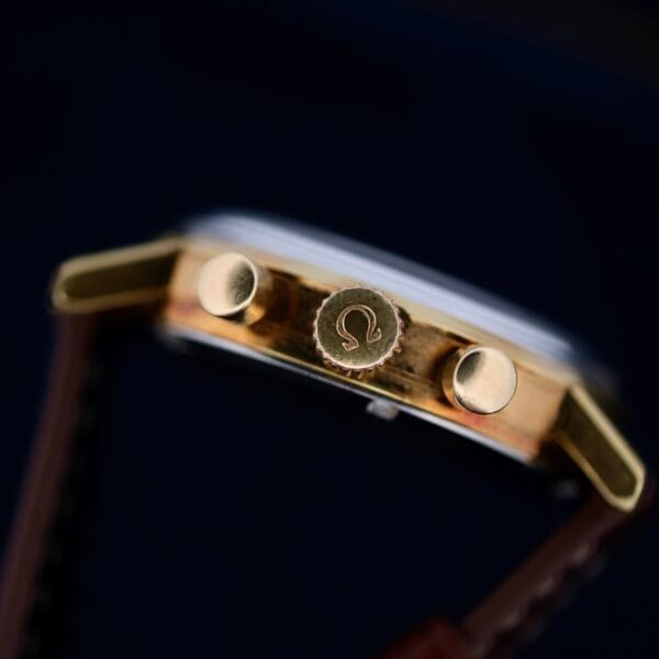 Close-up of the Omega Seamaster Chronograph Ref. 105.005 321 Caliber crown with an engraved logo, flanked by two push buttons, against a dark background. The watch features a partially visible brown leather strap.