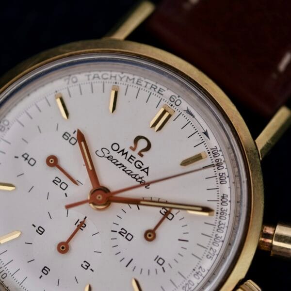 A close-up of the Omega Seamaster Chronograph Ref. 105.005 321 Caliber highlights its gold-tone bezel, white dial with tachymetre scale, and features gold hands, hour markers, three subdials, and a brown leather strap.