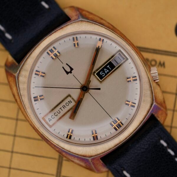 A vintage Bulova Accutron Day/Date watch with a round silver dial, gold hour markers, and a black leather strap features a SAT 11 display. Set on a beige background with partially visible text, it exudes mystery.
