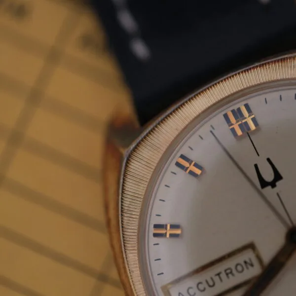 Close-up of a vintage Bulova Accutron Day/Date watch with an oxidized gold case. It features a white dial, gold-toned hour markers, and hands, exuding timeless elegance as it rests on a blurred patterned surface in the background.