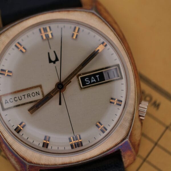 Close-up of the vintage Bulova Accutron Day/Date Gold Oxidized Case watch with a round gold face, displaying SAT and the date 11. It features a black leather strap set against a tan background with partial text.