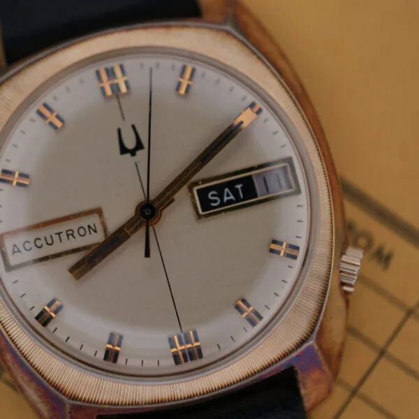 Close-up of the vintage Bulova Accutron Day/Date Gold Oxidized Case watch with a round gold face, displaying SAT and the date 11. It features a black leather strap set against a tan background with partial text.