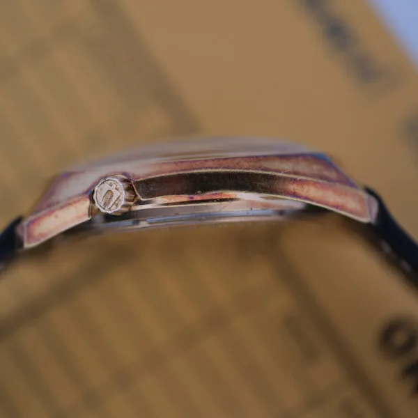 Close-up of a Bulova Accutron Day/Date Gold Oxidized Case wristwatch showing a worn gold-toned case and crown. Its set against a blurred background with possible text, and the watch band is partially visible.