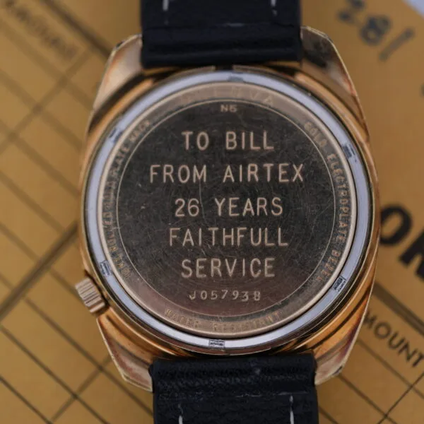 Close-up of the backplate from a Bulova Accutron Day/Date Gold Oxidized Case wristwatch, engraved with TO BILL FROM AIRTEX 26 YEARS FAITHFUL SERVICE and J057938. It features a black leather strap and rests on a brown surface with text.