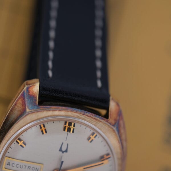 Close-up of a vintage Bulova Accutron Day/Date watch face with a gold oxidized case and black leather strap. It features yellow and black hour markers, an orange second hand, and the iconic Accutron branding prominently visible on the dial.