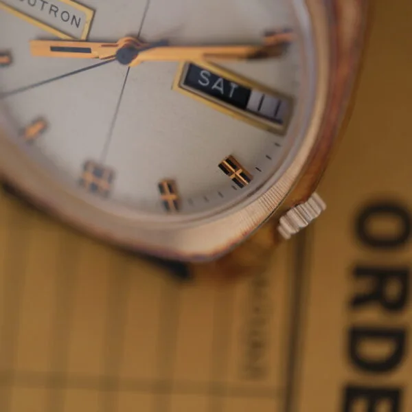 A close-up of a vintage Bulova Accutron Day/Date Gold Oxidized Case wristwatch shows SAT and 11. The backgrounds blurred paper features the word ORDER, enhancing the watchs timeless appeal.