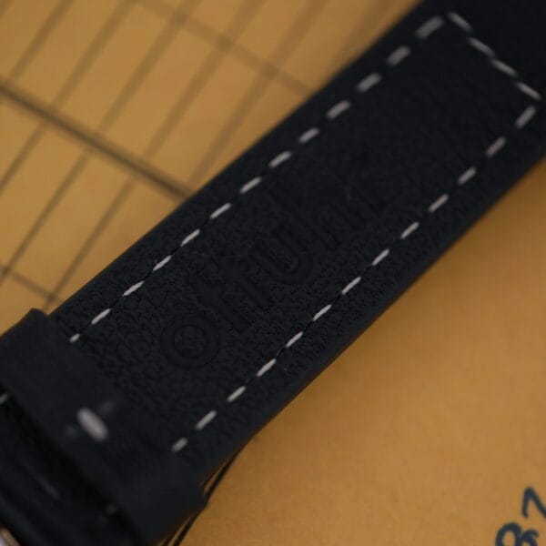 Close-up of a black leather watch strap with white stitching, embossed otfuhr, framed by a grid pattern and part of a brown envelope, evoking classic Bulova Accutron design elements from the Bulova Accutron Day/Date Gold Oxidized Case.