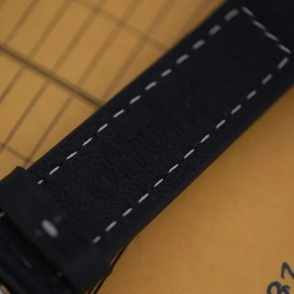 Close-up of a black leather watch strap with white stitching, embossed otfuhr, framed by a grid pattern and part of a brown envelope, evoking classic Bulova Accutron design elements from the Bulova Accutron Day/Date Gold Oxidized Case.