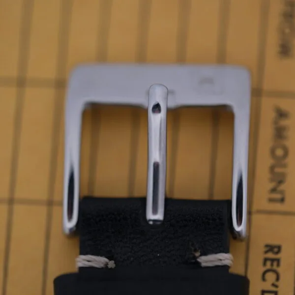 A close-up of a silver belt buckle on a black leather strap evokes the Bulova Accutron Day/Date Gold Oxidized Case watch design, resting gracefully on a surface with faint text and lines in the background.