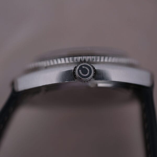 Close-up of a watch crown embossed with a stylized G. The Caravelle Automatic Ref 745 Diver “Jet Seconds” Super Waterproof boasts a sleek silver case with textured edges and a black strap. The blurred background highlights its side profile, emphasizing refined elegance.