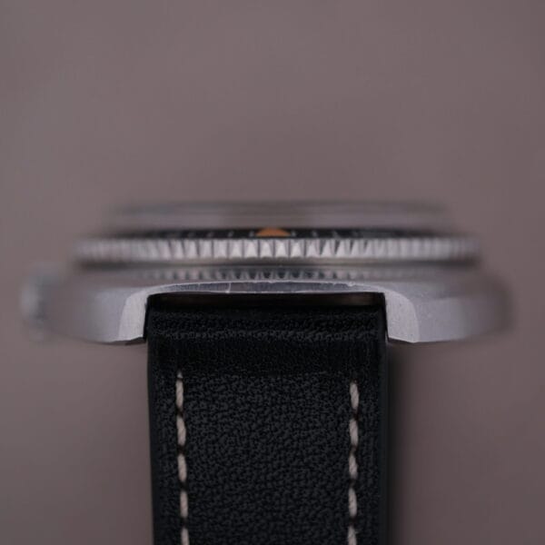 This close-up highlights the sleek silver metallic case and ridged edge of the Caravelle Automatic Ref 745 Diver “Jet Seconds” Super Waterproof wristwatch. Focused on its black leather strap with white stitching, set against a soft gray background, this piece exudes timeless elegance.
