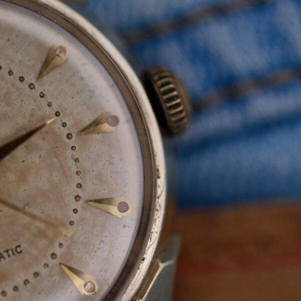 The Longines Automatic Caliber 19AS features a vintage watch face with a weathered dial, gold triangular hour markers, and twisted lugs. Its round metallic case and visible winding crown are set against a softly blurred blend of blue and brown fabric, enhancing its classic charm.
