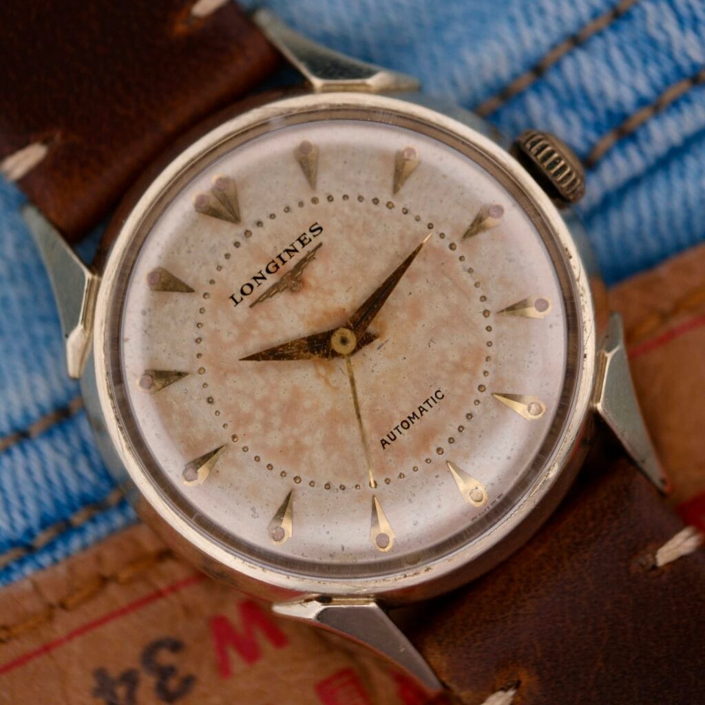 A Longines Automatic Caliber 19AS Warm Patina & Twisted Lugs wristwatch with a brown leather strap displayed on denim fabric.
